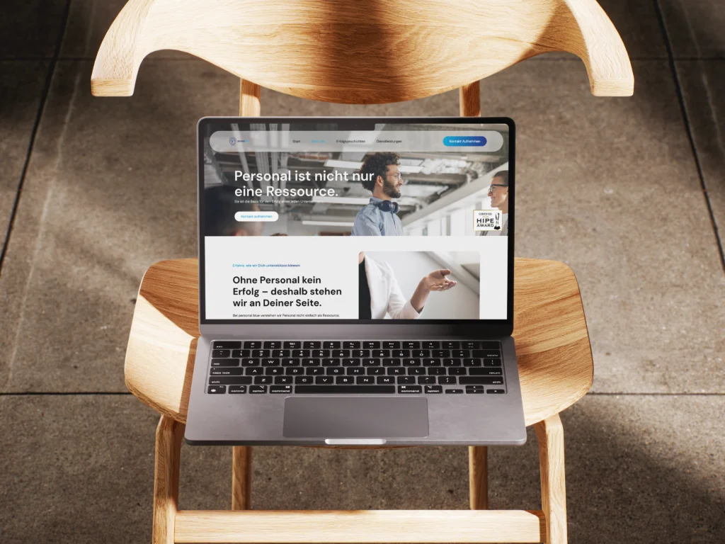 Laptop mit geöffneter Website der Personal Blue GmbH auf einem Holzstuhl, Startseite mit Slogan und Bild eines Gesprächs